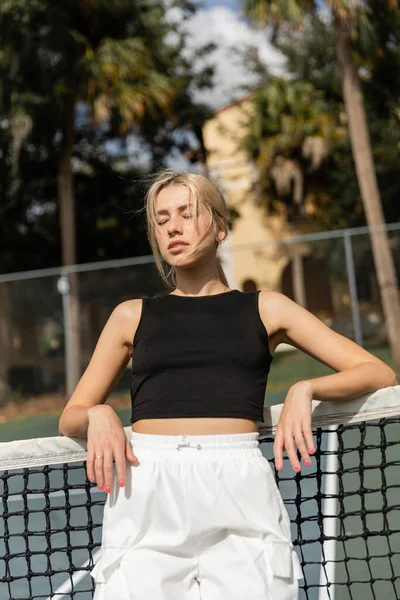 Mujer joven en camiseta negra y pantalones de carga blancos apoyados en la red en la cancha de tenis - foto de stock