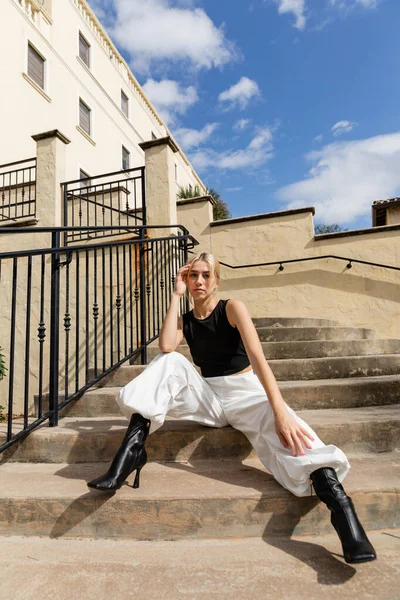 Pleine longueur de femme blonde en débardeur et pantalon cargo blanc assis sur les escaliers près du bâtiment à Miami — Photo de stock