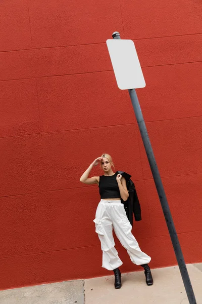 Повна довжина білявки у вантажних штанах стоїть зі шкіряною курткою біля червоної стіни в Маямі. — стокове фото