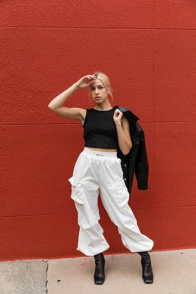 Piena lunghezza di donna elegante in pantaloni cargo in piedi con giacca camicia di pelle vicino alla parete rossa a Miami — Foto stock
