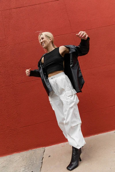 Piena lunghezza di donna allegra in pantaloni cargo e giacca di camicia di pelle in piedi vicino al muro rosso a Miami — Foto stock