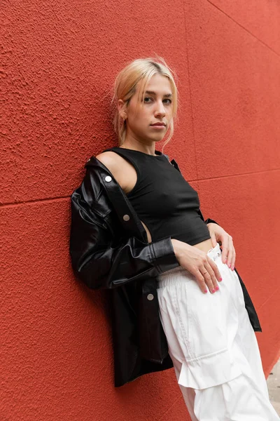 Donna bionda in pantaloni cargo e giacca di camicia di pelle in piedi vicino al muro rosso a Miami — Foto stock