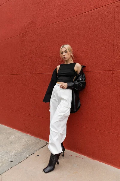 Piena lunghezza di donna bionda in pantaloni cargo e giacca di camicia di pelle in piedi vicino al muro rosso a Miami — Foto stock