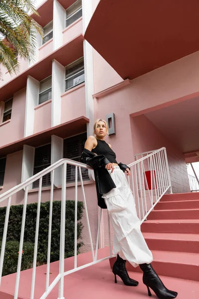 Longitud completa de mujer rubia en camiseta negra y pantalones de carga de pie en las escaleras de color rosa en Miami - foto de stock