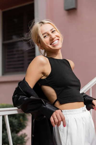 Cheerful blonde woman in black tank top and cargo pants standing near pink house in Miami — Stock Photo