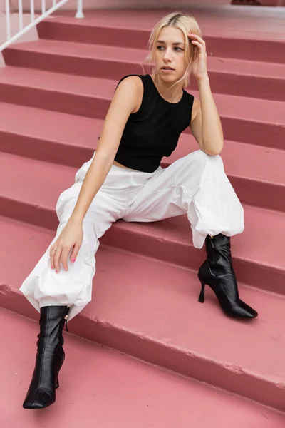 Pleine longueur de jeune femme blonde en débardeur noir et pantalon cargo assis sur des escaliers roses à Miami — Photo de stock