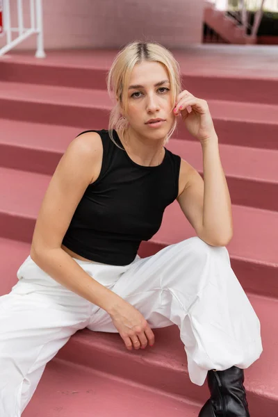 Blonde young woman in black tank top and cargo pants sitting on pink stairs in Miami — Stock Photo
