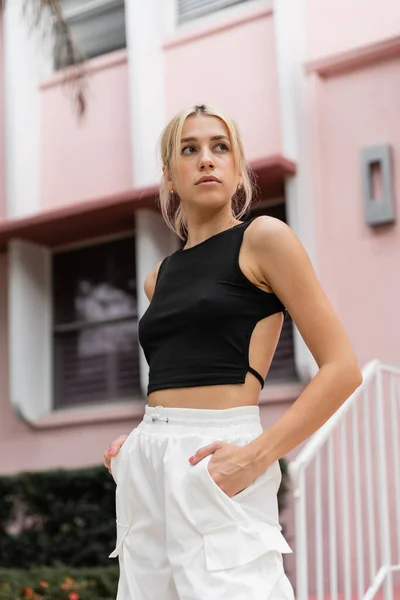 Giovane donna bionda in canotta nera e pantaloni cargo in piedi con le mani in tasca vicino alla casa rosa a Miami — Foto stock