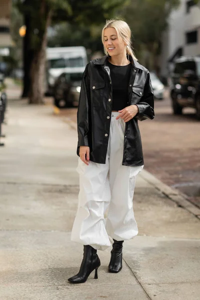 Comprimento total de mulher alegre em jaqueta de camisa de couro e calças de carga andando na rua urbana em Miami — Fotografia de Stock