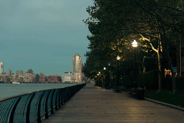 Светящиеся фонари возле деревьев на набережной реки Гудзон и вечерний городской пейзаж Манхэттена в Нью-Йорке — стоковое фото