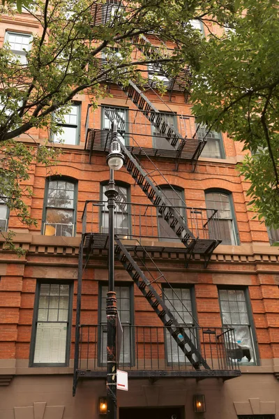 Brick dwelling building with metal balconies and fire escape stairs near lantern and trees in New York City - foto de stock
