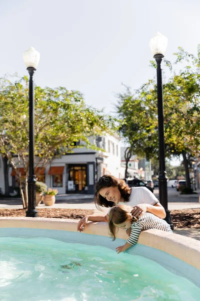 Mãe alegre olhando para a água na fonte perto da filha da criança na rua em Miami — Fotografia de Stock