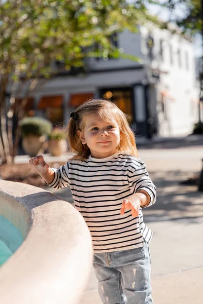 Маленька дівчинка в сорочці з довгими рукавами стоїть біля фонтану на вулиці в Маямі. — стокове фото