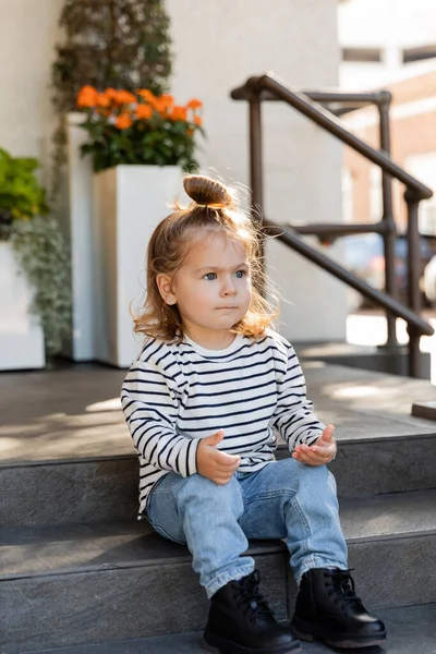 Toddler girl in long sleeve shirt and jeans sitting on stairs near house — Stock Photo