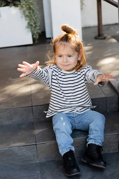 Kleinkind in Freizeitkleidung fordert Aufmerksamkeit und gestikuliert, während es auf einer Treppe in der Nähe des Hauses sitzt — Stockfoto