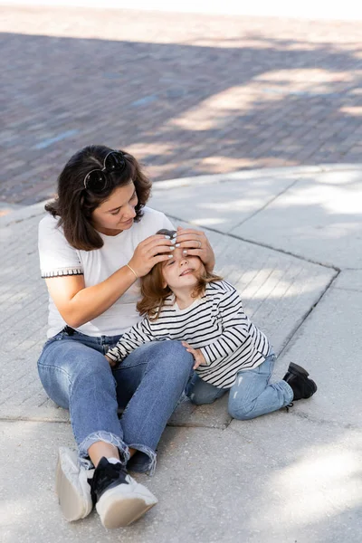 Brunette mother in jeans sitting with excited daughter in casual clothes on street in Miami — Stock Photo
