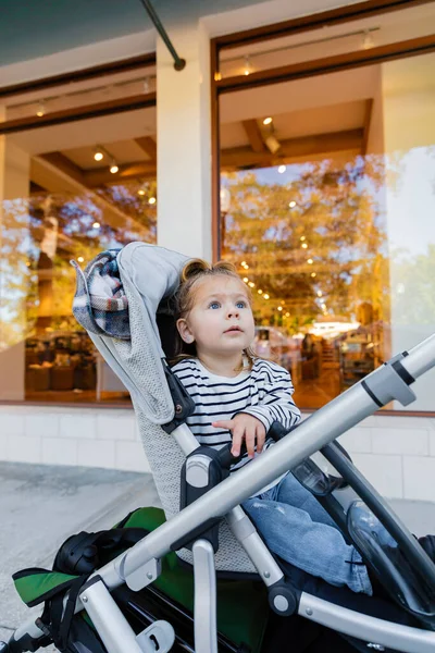 Toddler girl in striped long sleeve shirt sitting in baby stroller near showcase of shop in Miami — Stock Photo