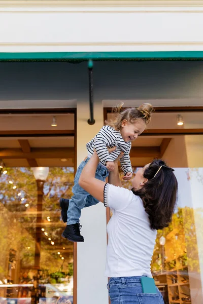 Madre levantamiento feliz bebé niña en rayas camisa de manga larga cerca de escaparate de tienda en Miami - foto de stock
