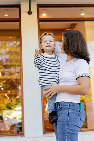Brunette mother holding in arms excited baby girl in striped long sleeve shirt near showcase of shop in Miami — Stock Photo