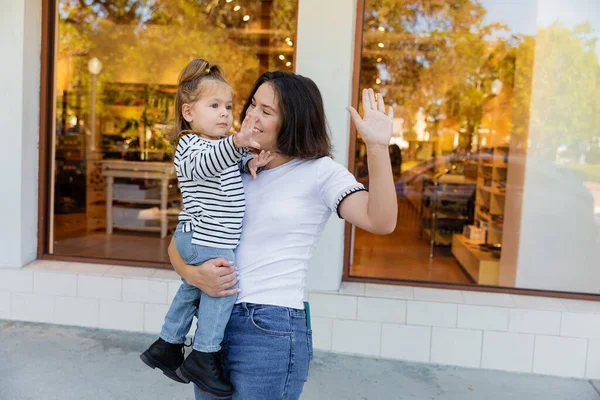 Happy mother holding in arms baby girl and gesturing together near showcase of shop in Miami — Stock Photo