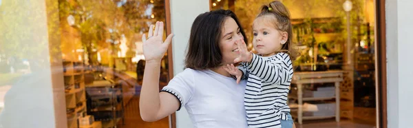 Felice madre in braccio bambina e gesticolare insieme vicino vetrina del negozio a Miami, banner — Foto stock
