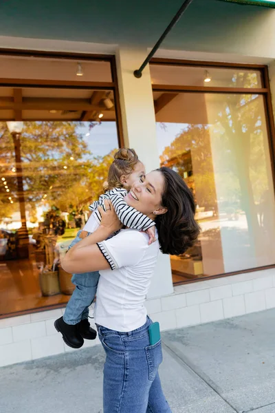 Glückliche Mutter umarmt Baby-Mädchen in gestreiftem Langarmshirt in der Nähe von Schaufenster des Geschäfts in Miami — Stockfoto