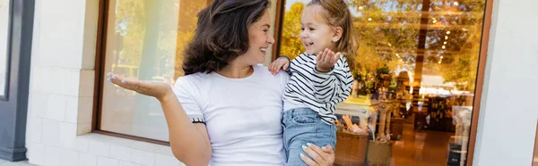 Cheerful mother holding in arms baby girl in striped long sleeve shirt near showcase of shop in Miami, banner — Stock Photo