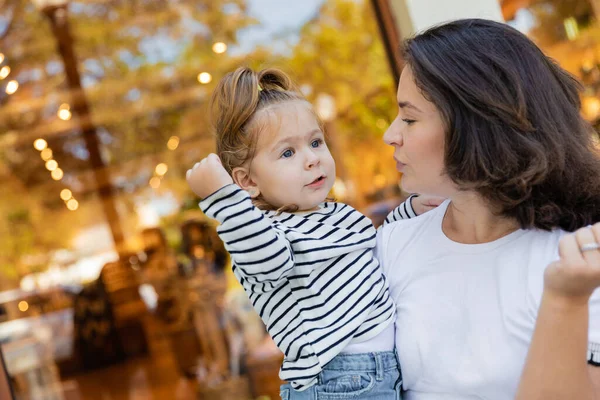 Brunette mother holding in arms baby girl in striped long sleeve shirt near showcase of shop in Miami — Stock Photo