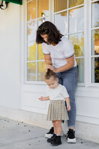 Мати в джинсах робить волосся дочки малюка в спідниці на ганку будинку — стокове фото