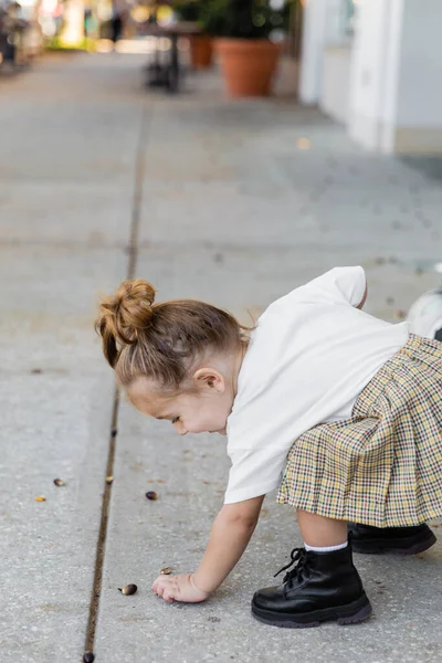 Happy toddler girl in skirt picking up acorn from ground on street in Miami — Stock Photo
