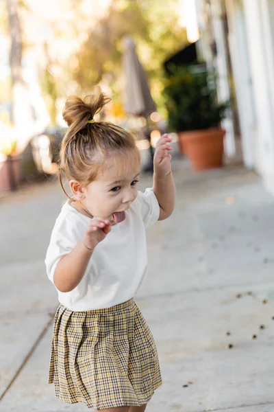 Дівчинка-малюк в спідниці і футболці, що стирчить язиком і жестикулює на відкритому повітрі — стокове фото