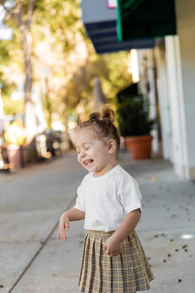 Feliz niña en falda y camiseta sonriendo con los ojos cerrados al aire libre - foto de stock