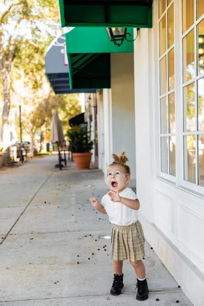 Налякана дівчинка-малюк в спідниці і футболці кричить біля будівлі — стокове фото