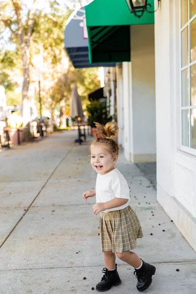 Повна довжина позитивної дівчинки-малюка в спідниці і футболці, що стоїть біля будівлі — стокове фото