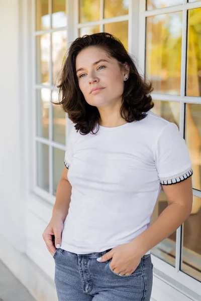 Ritratto di donna bruna in t-shirt bianca distogliendo lo sguardo mentre in piedi con le mani in tasca vicino a casa — Foto stock
