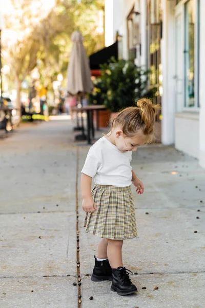 Pleine longueur de tout-petit fille en jupe et t-shirt blanc regardant le sol sur la rue à Miami — Photo de stock