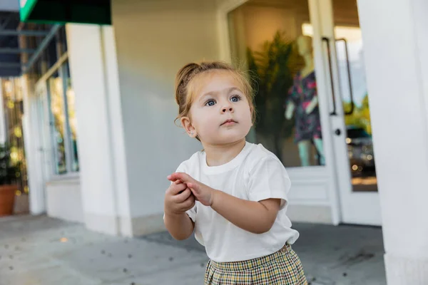 Маленькая девочка в белой футболке смотрит в сторону, стоя на улице — стоковое фото