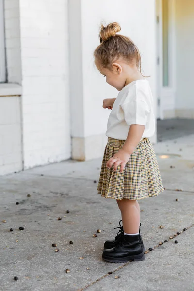 Longitud completa de niña en falda y camiseta blanca mirando bellotas en el suelo - foto de stock