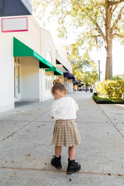 Longitud completa de la niña en falda y camiseta blanca mirando bellotas en el suelo en Miami - foto de stock