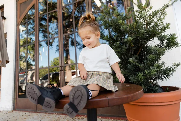 Comprimento total da menina da criança sorridente em saia e camiseta branca sentada na mesa de bistrô perto de café ao ar livre em Miami — Fotografia de Stock