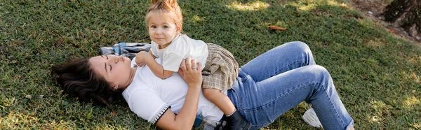 Morena madre haciendo pucheros labios y acostado en la hierba con niño niña, pancarta - foto de stock