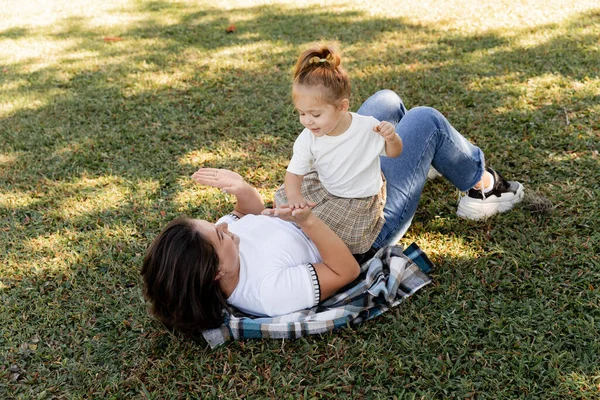 Веселая мать, лежащая на зеленой лужайке и играющая в пирожные с веселой маленькой девочкой в парке Майами — стоковое фото