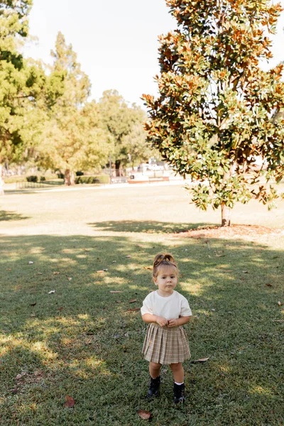 Pleine longueur de tout-petit fille en jupe et t-shirt blanc debout dans le parc avec des arbres verts — Photo de stock