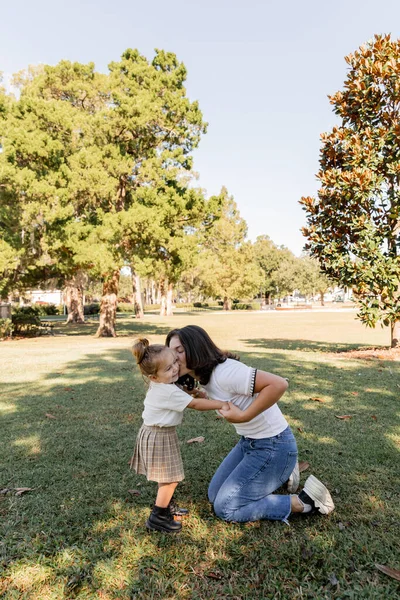 Brunette mother standing on knees and kissing cheek of toddler daughter in park of Miami — Stock Photo