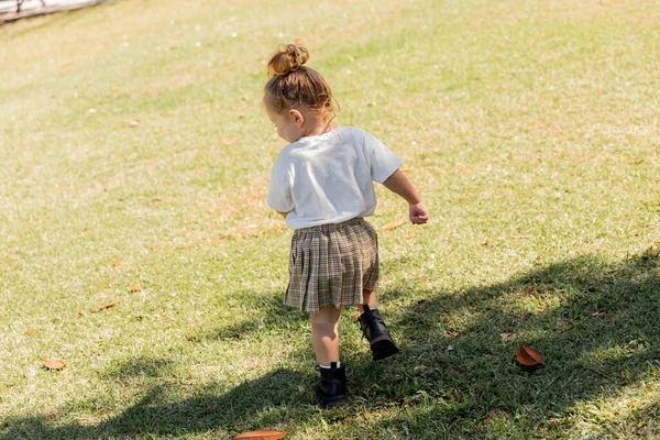 Back view of toddler girl in white t-shirt and skirt walking in boots on green grass — Stock Photo