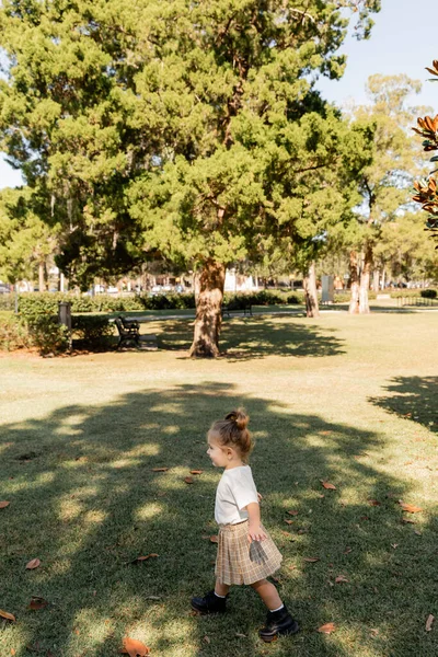 Joyeuse petite fille en t-shirt blanc et jupe jouant dans un parc vert — Photo de stock