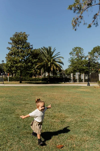 Позитивна дівчинка-малюк стоїть з простягнутими руками, граючи в зеленому парку — стокове фото