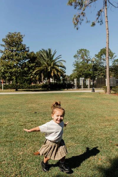Радісна дівчинка-малюк стоїть з витягнутими руками, граючи в зеленому парку — стокове фото