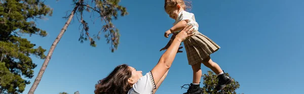 Feliz madre levantando hija pequeña contra árboles verdes y cielo despejado, bandera - foto de stock