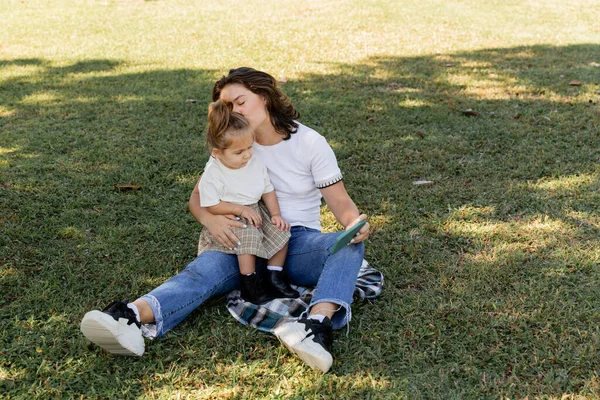 Madre baciare la bambina mentre tiene smartphone e seduto sul prato — Foto stock
